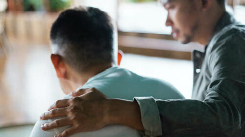 A man's slumped back with another man comforting him with his arm around his shoulder.