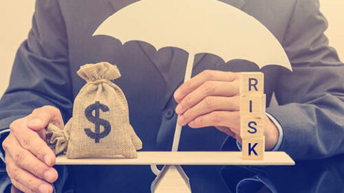 A man in a suit balancing a bag of money and blocks that say risk with an umbrella over them.