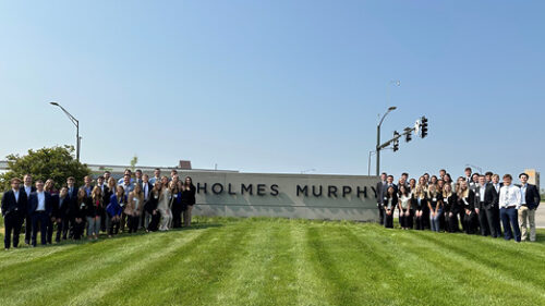 A group photo of the class of 2023 Holmes Murphy interns in front of the Holmes Murphy sign on a green lawn.