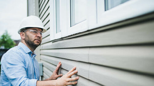 A man in a hardhat inspecting siding of a house.